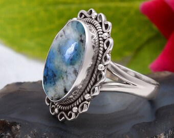 May include: A silver ring featuring a large, oval kyanite stone with blue and white patterns. The stone is set in a decorative, ornate bezel. The ring is displayed on a dark, textured surface, with a rose and green leaf in the background.