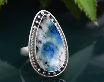 May include: A silver ring featuring a large, teardrop-shaped stone. The stone has a white base with blue and gray patterns. The ring's setting has a beaded border and a twisted rope design. The ring is displayed on a reflective surface.