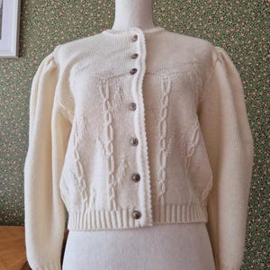 Könnte beinhalten: Eine cremefarbene Strickjacke mit langen Puffärmeln, rundem Halsausschnitt und einer Knopfleiste mit metallischen Knöpfen. Das Kleidungsstück zeigt ein strukturiertes Zopfmuster auf der Vorderseite sowie gerippte Bündchen und Saum.
