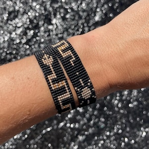 May include: Two black beaded bracelets with gold and silver accents. The bracelets are stacked on a person's wrist.