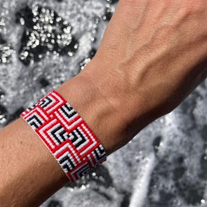 Può includere: Un braccialetto intrecciato rosso, nero e bianco con un motivo geometrico. Il braccialetto è indossato al polso.