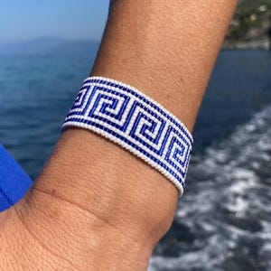 Peut inclure: Un bracelet en perles blanches et bleues avec un motif de clé grecque. Le bracelet est porté au poignet.