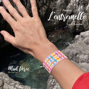 Peut inclure: Un bracelet tissé avec une base rose et un motif coloré de bleu, jaune, orange et blanc. Le bracelet est sur un poignet, avec le texte "Pattern L'entremelle Loom Bracelet" et "Madi Perec Jewelry Pop!" dans l'image.