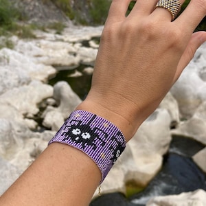 May include: Purple beaded bracelet with a black and white pixelated design of a cat's face. The bracelet has gold beads and a gold chain.