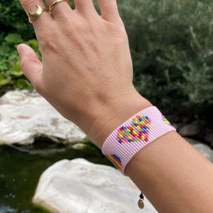 May include: A pink woven bracelet featuring a colourful heart design. The bracelet is made with small beads and has a gold charm at the end.