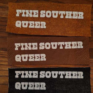 May include: Three rectangular patches in shades of brown and black, each bearing the text "FINE SOUTHER QUEER" in white, glittery lettering. The patches are made of different materials, including felt and leather.