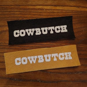 May include: Two rectangular patches with the word "COWBOUTCH" in glittery white letters. One patch is black, the other is tan. The patches are displayed on a wooden surface.