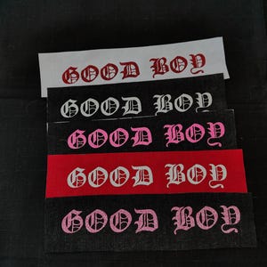 May include: Five fabric patches with the words "GOOD BOY" in a gothic font. The patches are in different colors, including white, pink, and red.