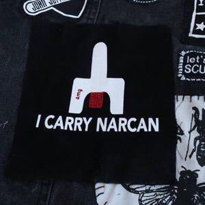 May include: Black square patch with white text reading "I CARRY NARCAN." The patch features a white graphic with a red glitter accent and the text "4mg." The patch is sewn onto a denim jacket.