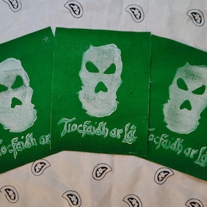 May include: Three green fabric squares with a white skull design and the text "Tiocfaidh ar Lá". The squares are arranged on a white paisley patterned background.