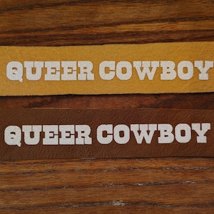 May include: Two rectangular patches with the text "QUEER COWBOY" in white block letters. One patch is mustard yellow, the other is brown. The patches are on a wooden surface.