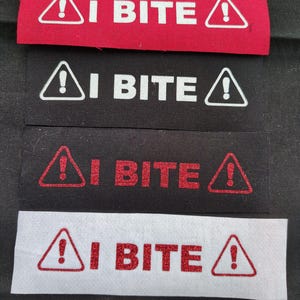 May include: Four fabric patches with the text "I BITE" in white and red lettering on a black and white background. The patches have a red exclamation point inside a white triangle.