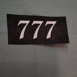 May include: A black rectangular patch with the number "777" in white, glittery font. The patch is set against a teal fabric background. The numbers are bold and stylized.