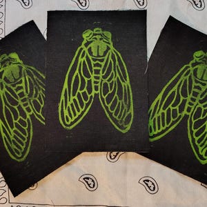 May include: Three black prints featuring a green cicada insect design. The cicadas are depicted with detailed wing patterns. The prints are arranged on a white and black paisley patterned fabric.