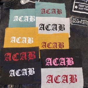 May include: A collection of rectangular patches with the letters "ACAB" in various colors and fonts. The patches are in shades of red, pink, white, and black, with some featuring glitter. The patches are displayed on a dark denim jacket.