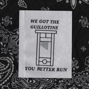 May include: A white fabric patch with the text "WE GOT THE GUILLOTINE YOU BETTER RUN" in black. A guillotine illustration with a silver blade is centered on the patch. The background is a black and white paisley pattern.