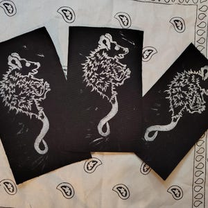 May include: Three black art prints featuring a white linocut of a possum. The possum is depicted with a fluffy body and a long, curved tail. The prints are arranged on a white paisley patterned fabric.