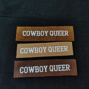 May include: Three brown leather patches with the text "COWBOY QUEER" in white lettering.