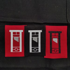 May include: Three fabric patches featuring a guillotine design. Two patches have a red background, one with a white guillotine and the other with a black guillotine. The third patch has a black background with a white guillotine.