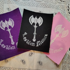 May include: Three patches in purple, black, and pink, each featuring a white axe design and the words "Lesbian Menace" in a gothic font. The patches are arranged on a paisley patterned fabric.