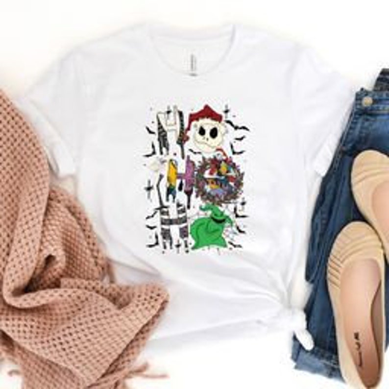 May include: White t-shirt featuring a graphic design with characters from The Nightmare Before Christmas. The design includes Jack Skellington, Sally, and Zero, with the word "HALLOWEEN" incorporated into the artwork. The shirt is paired with denim jeans and beige flats.