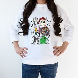 May include: White sweatshirt featuring a colorful graphic of characters from "The Nightmare Before Christmas." The design includes Jack Skellington, Sally, Oogie Boogie, and other elements from the movie. The sweatshirt has long sleeves and a crew neck.