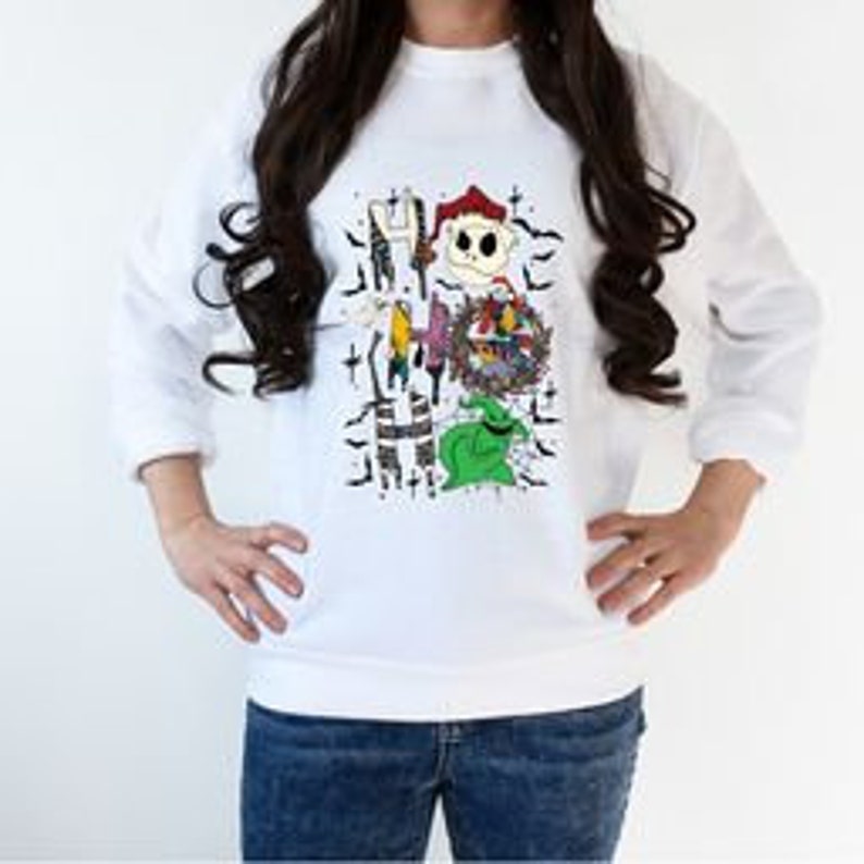 May include: White sweatshirt featuring a colorful graphic of characters from "The Nightmare Before Christmas." The design includes Jack Skellington, Sally, Oogie Boogie, and other elements from the movie. The sweatshirt has long sleeves and a crew neck.