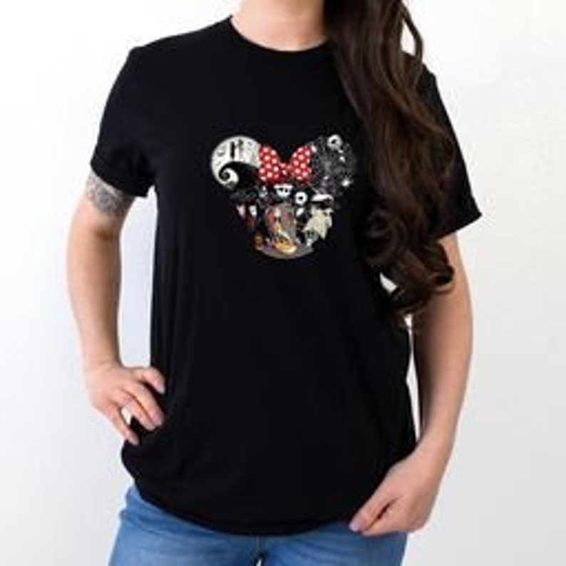 May include: Black crew neck t-shirt with a graphic of a Mickey Mouse head and a red bow. The graphic is filled with characters and imagery from The Nightmare Before Christmas. The shirt is worn by a person.