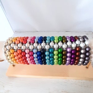 May include: A display of beaded bracelets in a rainbow of colors. Each bracelet is made of round, pearl-like beads and silver-colored spacers. The bracelets are arranged on a light-colored wooden display stand.