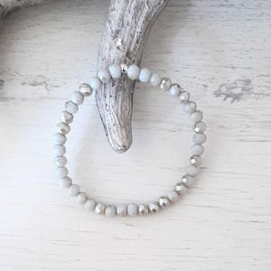 Pode incluir: Uma pulseira de contas cinza claro com detalhes prateados. A pulseira é feita de pequenas contas redondas enfiadas, criando um acessório delicado e elegante. A pulseira é exibida em uma superfície de madeira branca.
