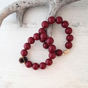 Könnte beinhalten: Zwei weinrote Perlenarmbänder. Ein Armband hat einen goldenen Verschluss mit klaren Kristallen, das andere einen Leopardenmuster-Akzent. Die Armbänder werden auf einer weißen Holzoberfläche mit einem dekorativen Geweih präsentiert.