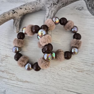 Könnte beinhalten: Zwei Armbänder mit Perlen. Jedes Armband hat dunkelbraune Perlen, irisierende Perlen und flauschige, hellbraune Pompons. Die Armbänder werden auf einer hellen Holzoberfläche mit einem dekorativen Ast präsentiert.