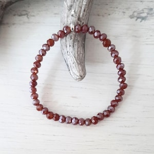 Könnte beinhalten: Ein Stretch-Armband aus kleinen, facettierten, rotbraunen Perlen. Die Perlen sind zu einer kreisförmigen Form aufgereiht. Das Armband wird auf einer hellen Holzoberfläche mit einem Stück Treibholz präsentiert.
