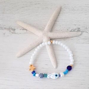 May include: White beaded bracelet with gold accents and a fish charm. The bracelet features a gold starfish charm, blue and white beads, and a fish charm with blue polka dots. A white starfish is in the background.