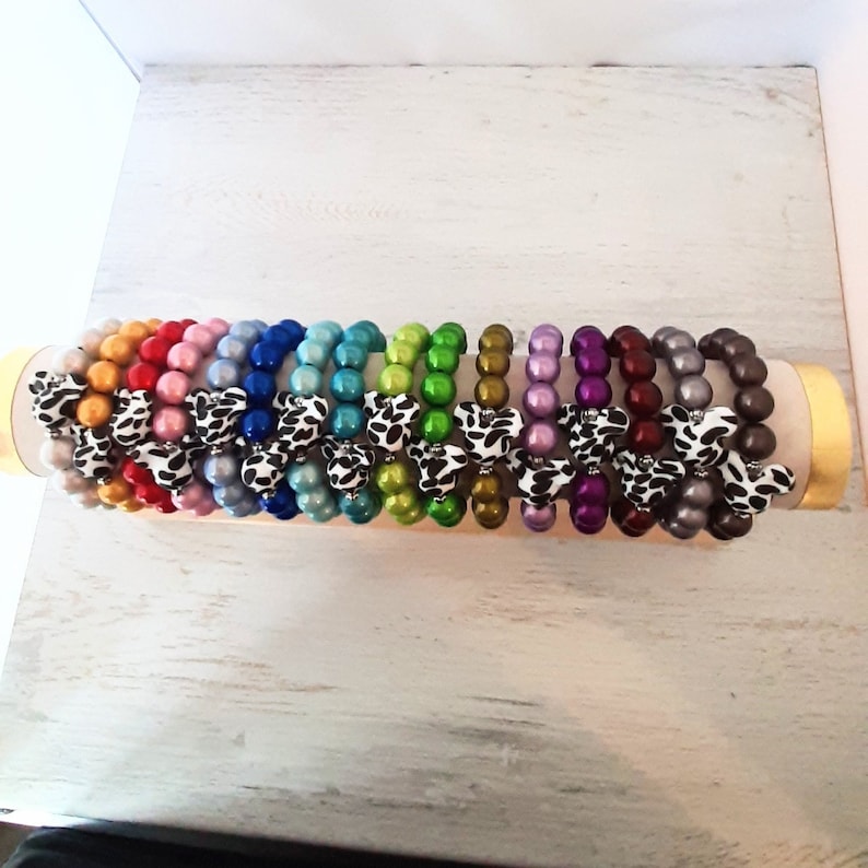 May include: A set of 14 colorful beaded bracelets with a cow print bow accent. The bracelets are displayed on a wooden stand.