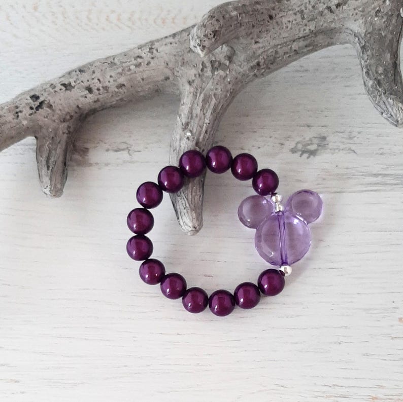 May include: A purple beaded bracelet with a purple Mickey Mouse charm. The bracelet is made with small, round beads and is strung on a silver chain.