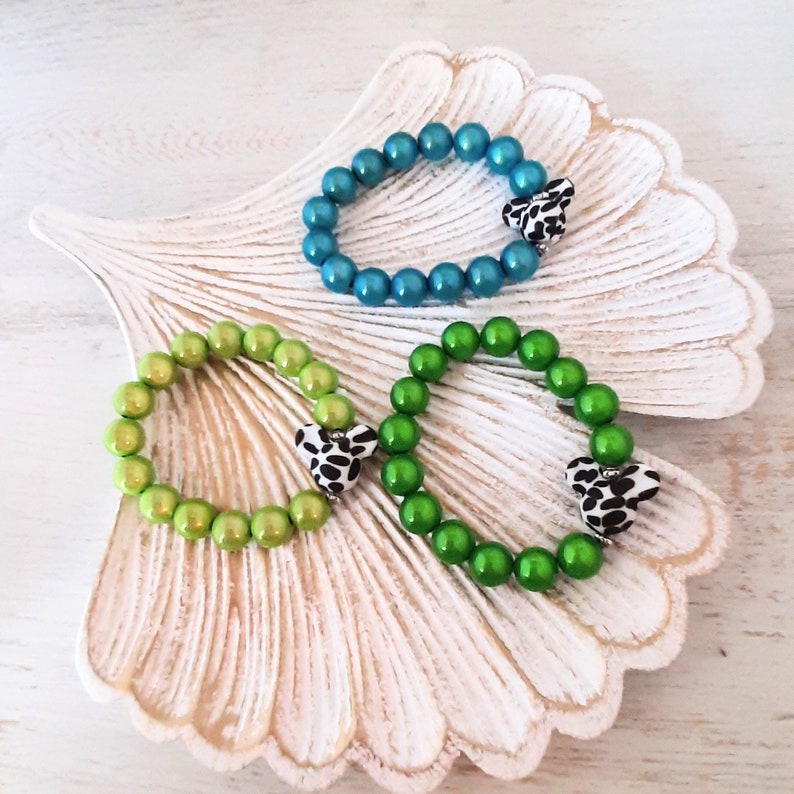 May include: Three beaded bracelets with heart-shaped charms. The bracelets are green, blue, and light green. The charms are black and white with a spotted pattern.