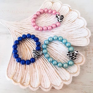 May include: Three bracelets made of colorful pearl beads with a black and white animal print charm on each. The bracelets are displayed on a white seashell-shaped surface.