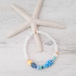 May include: A white beaded bracelet with a blue fish charm. The fish has white polka dots and is accompanied by blue and turquoise beads. Gold-colored accents are present on the bracelet.