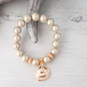 Puede incluir: Un brazalete de perlas beige con un dije de concha dorada. El brazalete está hecho de perlas redondas y tiene un cierre dorado.