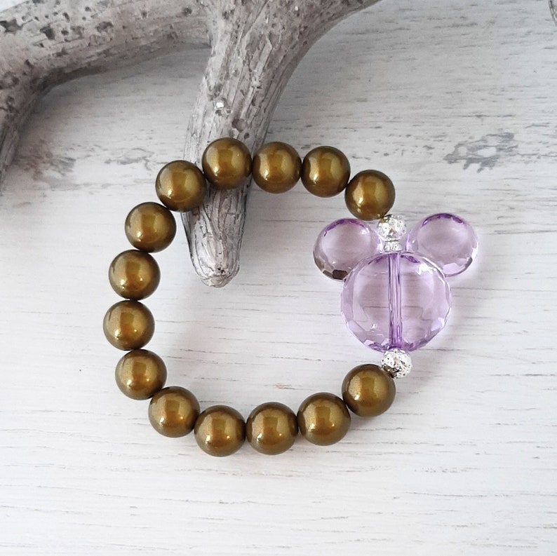May include: A brown beaded bracelet with a purple Mickey Mouse charm. The bracelet is made of round beads and the charm is made of faceted glass.