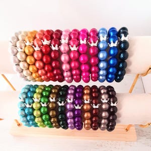 May include: A set of 20 colourful bracelets with silver crown charms. Each bracelet is made of round, shiny beads. The bracelets are arranged in a rainbow pattern on a wooden display stand.