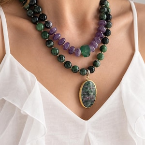 May include: A multi-strand beaded necklace featuring dark green and black mottled stone beads, purple amethyst-coloured beads, and a central green stone accent. A large oval pendant with a mottled green and red stone set in a gold-toned frame.