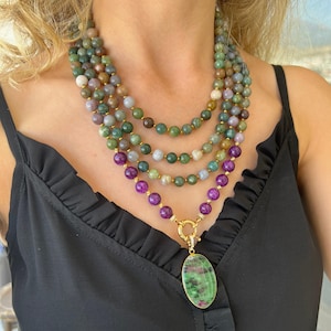 May include: A multi-strand necklace featuring green, purple and brown beads. The necklace has a large oval pendant with a green and pink stone.
