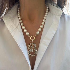 May include: A white pearl necklace with a gold charm in the shape of the number "3" with small diamonds. The charm is attached to a gold ring.