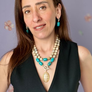 May include: A woman wearing a two-strand necklace with turquoise and beige beads, a gold pendant, and matching turquoise earrings. The pendant is oval-shaped with a striped design. The woman is wearing a black top.