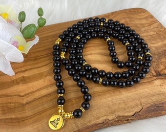 May include: Black beaded mala necklace with gold accents and a gold Buddha charm. The necklace is laid out on a wooden surface.