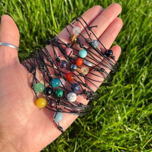 May include: A collection of gemstone bead bracelets with black cords. The beads are various colors, including green, blue, red, and yellow. The bracelets are displayed on a hand against a backdrop of green grass.