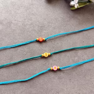 May include: Three turquoise cord bracelets, each featuring a single round letter bead flanked by two small gold-toned metallic beads. The top bead is red with an S, the middle is yellow with an S, and the bottom is pink with an A.