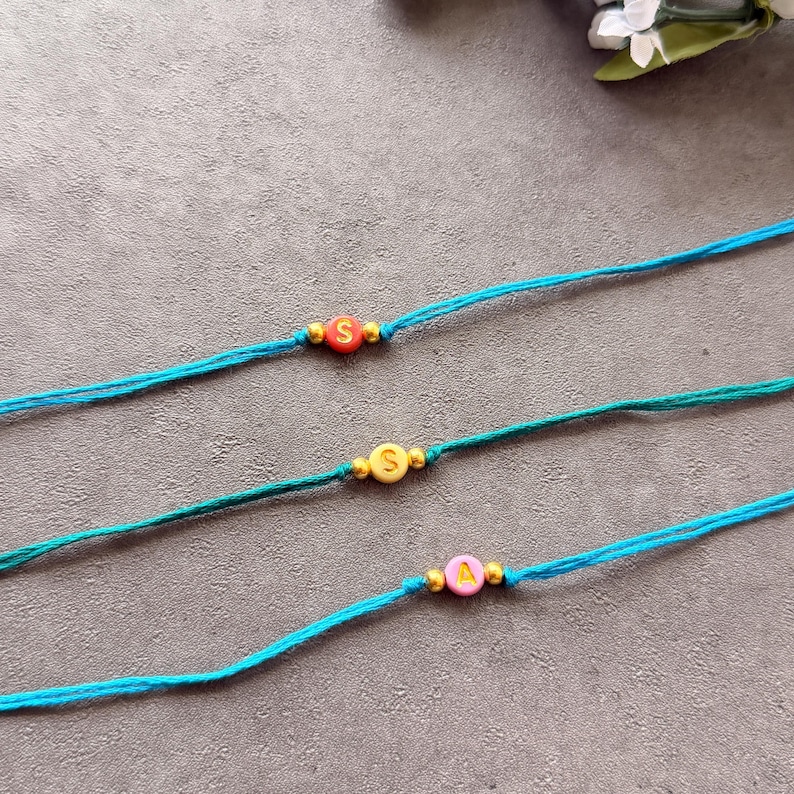 May include: Three turquoise cord bracelets, each featuring a single round letter bead flanked by two small gold-toned metallic beads. The top bead is red with an S, the middle is yellow with an S, and the bottom is pink with an A.